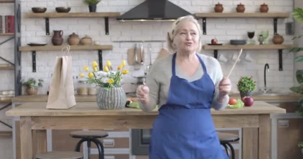 Mulher caucasiana sênior alegre dançando com utilitários de cozinha na cozinha moderna. Retrato de aposentado de meia-idade alegre se divertindo em casa com pensão. Felicidade, estilo de vida, alegria. Cinema 4k ProRes HQ . — Vídeo de Stock