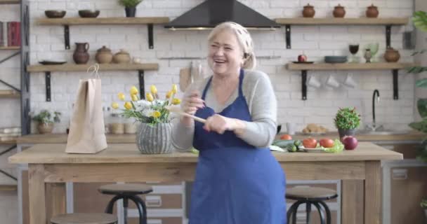 Retrato de reformado feminino alegre dançando e girando com utensílios de cozinha dentro de casa. Sorrindo caucasiano cruzando as mãos e olhando para a câmera. Diversão, alegria, lazer, estilo de vida. Cinema 4k ProRes HQ . — Vídeo de Stock