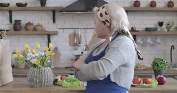 Portrait de femme caucasienne souriante positive regardant en arrière à la cuisine moderne et se tournant vers la caméra. Femme retraitée insouciante posant avec ustensile de cuisine à l'intérieur. Siège social Cinema 4k ProRes . — Video