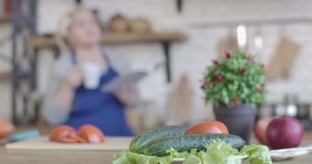 Gros plan de légumes frais couchés sur la table dans la cuisine en tant que femme floue buvant du café et lisant un livre de recettes en arrière-plan. Salade de cuisine pour retraités d'âge moyen le matin. Siège social Cinema 4k ProRes — Video