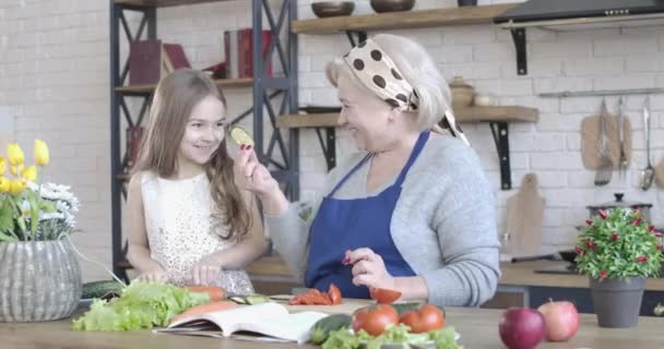 Mutfakta doğranmış salatalık tadı alan pozitif beyaz büyükanne ve torunun portresi. Şirin küçük kız son sınıftaki bayana evde salata pişirmesi için yardım ediyor. Birlik, yaşam tarzı. Sinema 4k ProRes Merkezi. — Stok video