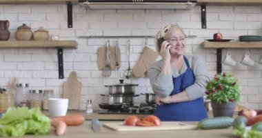 Evde, mutfakta telefonda konuşan yaşlı, pozitif bir emekli. Olgun, neşeli, beyaz bir kadının portresi. Kapalı mekanda durmuş sohbet ediyor. Yemek pişirme, yaşam tarzı, boş zaman. Sinema 4k ProRes Merkezi.