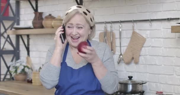 Portrait of cheerful female retiree eating apple and talking on the phone. Joyful blond Caucasian woman laughing as chatting in kitchen at home. Leisure, retirement, lifestyle. Cinema 4k ProRes HQ. — Stock Video