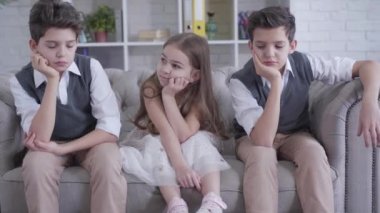 Kafkasyalı ikiz kardeşler, neşeli tatlı kızın her iki tarafında kanepede oturuyorlar. Evde dinlenen sıkılmış çocukların portresi. Arkadaşlık, çocukluk, rahatlık, yaşam tarzı..