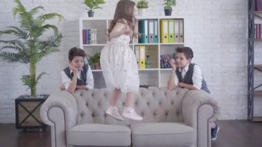 Yorgun ikiz kardeşler kanepenin iki tarafında durup neşeli kız kardeşlerinin kanepeye zıplamasını izliyorlar. Kardeşlerin evde daha genç esmer bir kızla vakit geçirmesinden bıktım. Yaşam tarzı, birlik, aile.