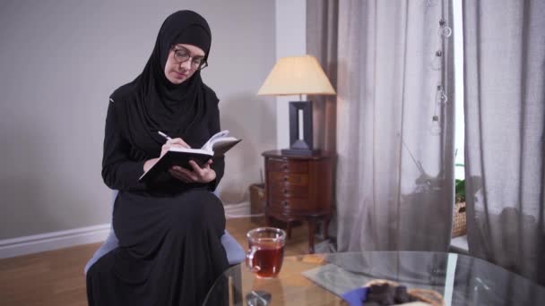 Konsentrasi wanita Muslim dengan kacamata tertulis di rumah. Sukses penulis perempuan modern dalam jilbab menciptakan best seller baru. Kreativitas, gaya hidup, penulis . — Stok Video
