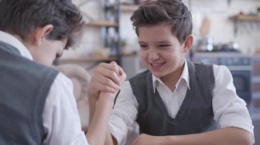 Bilek güreşinde ikiz kardeşiyle yarışan neşeli beyaz çocuğun portresi. Gülümseyen özdeş çocuklar evde eğleniyorlar. Eğlence, eğlence, yaşam tarzı, birlik.