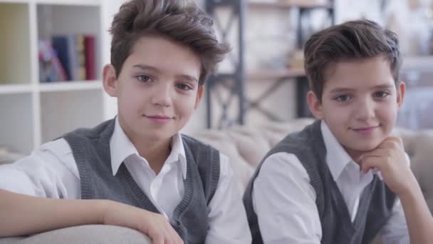 Gros plan portrait de beaux petits frères jumeaux caucasiens regardant la caméra et souriant. Garçons bruns positifs dans des vêtements élégants similaires assis sur le canapé à l'intérieur. Mode de vie, beauté, bonheur . — Video
