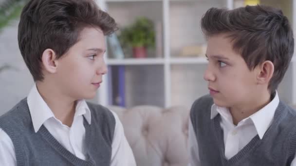 Close-up side view of two identical twin brothers looking at each other and smiling. Stylish brunette Caucasian boys in similar elegant clothes posing indoors. Happiness, unity, lifestyle. — Stock Video