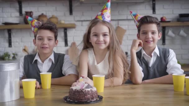 Cámara lenta, dos chicos gemelos caucásicos y linda chica en sombreros de fiesta sentado en la mesa con pastel de cumpleaños, mirando a la cámara, hablando y sonriendo. Cámara acercándose a los niños felices celebrando . — Vídeo de stock