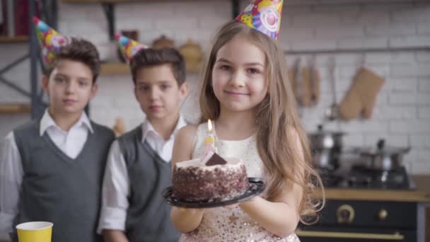 Retrato de una pequeña chica caucásica positiva en sombrero de fiesta sosteniendo pastel de cumpleaños y mirando a la cámara. Sus hermanos gemelos de pie en el fondo. Lento. Unidad, felicidad, celebración, estilo de vida . — Vídeos de Stock