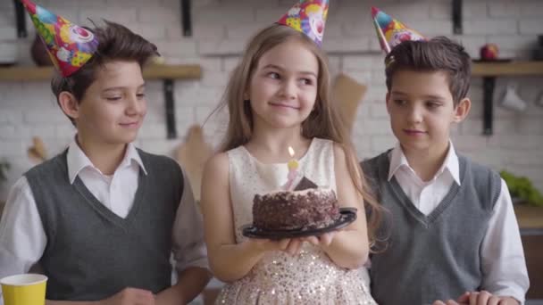 Lento de chica caucásica bastante feliz sosteniendo pastel de cumpleaños y soplando velas. Hermanos gemelos de pie a ambos lados aplaudiendo. Felicidad, alegría, estilo de vida, celebración. Movimiento lento . — Vídeos de Stock