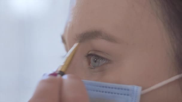 Vista lateral de cerca de la joven mujer caucásica en máscara protectora aplicando maquillaje. Chica usando lápiz de cejas durante la cuarentena. Belleza, pandemia, Covid-19 . — Vídeos de Stock