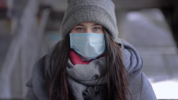 Kışlık giysiler içindeki esmer gri gözlü kız portresi ve kameraya bakan koruyucu maske. Covid-19 karantinası sırasında dışarıda poz veren beyaz bir kadın. Sağlık, güvenlik, yaşam tarzı, izolasyon — Stok video