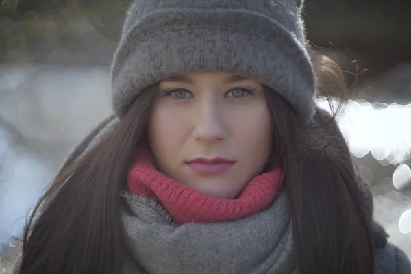 Close-up face of serious Caucasian girl in warm hat and scarf looking at camera. Portrait of young pretty woman outdoors.