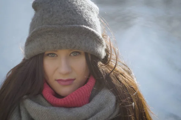 Close-up face of brunette woman in warm grey hat at the background of water. Portrait of pretty Caucasian girl standing outdoors at river bank or lake shore.