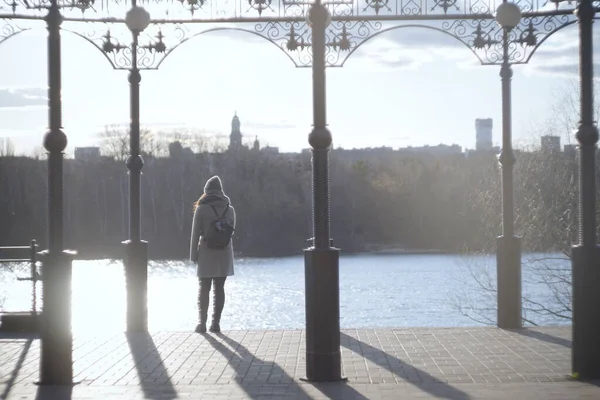 Tunnistamaton nuori nainen seisoo järven rannalla kaupungin puistossa. Takaisin näkymä tyttö lämpimissä vaatteissa reppu lepää kaupunkien vauhtia elämän. Elämäntapa, vapaa-aika, luonto . — kuvapankkivalokuva