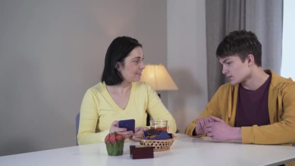 Jeune femme blanche brune assise à la table et utilisant le téléphone comme adolescent pour annoncer de mauvaises nouvelles. Fils apportant des informations choquantes à la mère. Drame, problème, trouble . — Video