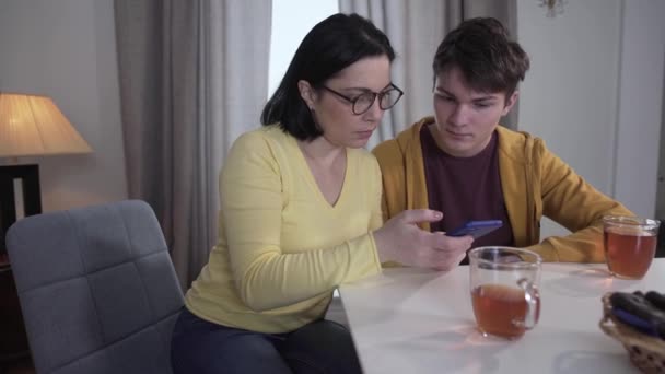 Tonåring brunett vit pojke lära vuxna mor i glasögon för att använda smartphone. Patient son hjälper kvinnan att hantera elektronik. Enhet, stöd, livsstil, vård. — Stockvideo