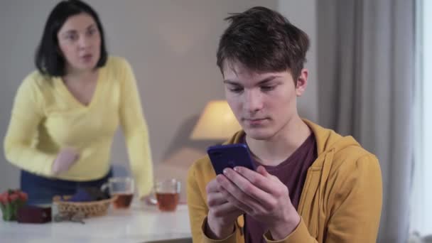 Close-up portret van brunette Kaukasische tiener jongen spelen van games op de smartphone als zijn moeder hem uitschelden op de achtergrond. Angstige wazige vrouw gebaren emotioneel tonen apparaat verslaving. — Stockvideo