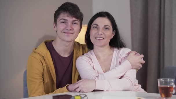 Meio tiro de filho e mãe caucasiano feliz olhando para a câmera e sorrindo como sentado na mesa dentro de casa. Adolescente beijando a mãe na bochecha e abraçando os pais. Alegria, felicidade, estilo de vida, unidade . — Vídeo de Stock