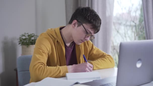 Πορτρέτο του συγκεντρωμένου Καυκάσιου έφηβου με γυαλιά που διαβάζει. Έξυπνος φοιτητής που διαβάζει από το λάπτοπ και γράφει στο βιβλίο εργασίας. Νοημοσύνη, εκπαίδευση, τρόπος ζωής. — Αρχείο Βίντεο