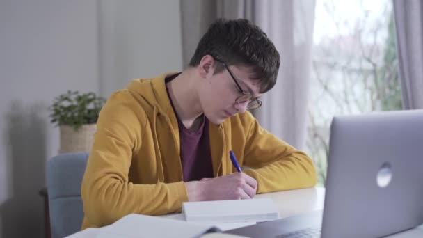 Retrato de un adolescente caucásico concentrado con anteojos haciendo la tarea. Estudiante universitario inteligente leyendo desde el portátil y escribiendo en el libro de trabajo. Inteligencia, educación, estilo de vida . — Vídeo de stock