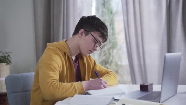 Die Kamera bewegt sich von links nach rechts um den jungen Kaukasier mit Brille und Laptop. Intelligenter Nerd-Student macht Hausaufgaben am Tisch. Bildung, Intelligenz, Lebensstil. — Stockvideo