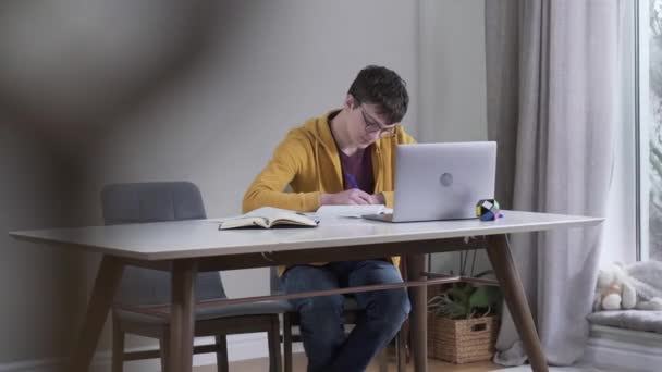 Largo tiro de chico caucásico inteligente en gafas mirando la pantalla del ordenador portátil y la escritura. Estudiante adolescente confiado estudiando duro en interiores. Educación, estilo de vida, inteligencia. Disparos detrás de balaustres . — Vídeos de Stock