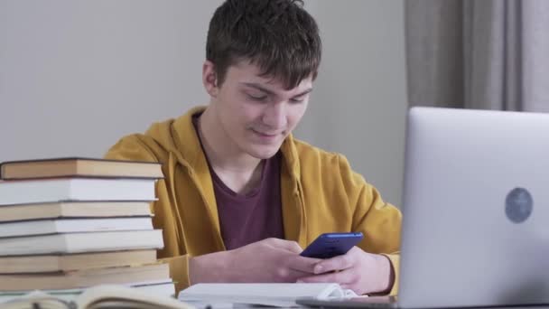 Close-up portrait of brunette Caucasian boy sitting at the table with books and laptop and using smartphone. Smiling teenager typing messages while studying. Education, lifestyle, modern technologies. — Stock Video