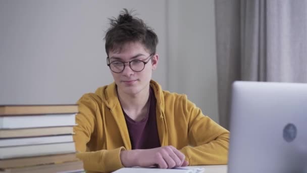 Portrait of exhausted Caucasian boy in eyeglasses looking at stack of books and laptop on the table and signing. Tired teenager studying hard indoors. Intelligence, lifestyle, education, overload. — Stock Video