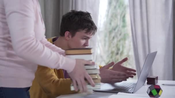 Side view of young Caucasian boy gaming as unrecognizable woman putting stack of books on table and slapping his head. Strict mother forcing son to study, lazy teenager continues game. Behavior. — Stock Video