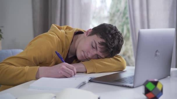 Slimme blanke tienerjongen die in slaap valt als schrijvend in een werkboek. Portret van een uitgeputte student die binnen hard studeert. Vermoeidheid, overwerk, onderwijs, levensstijl. — Stockvideo