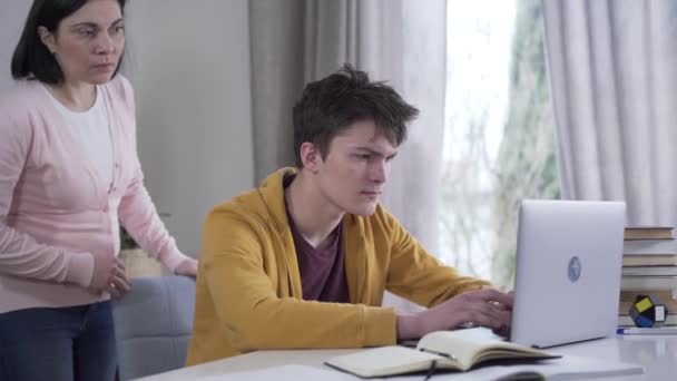 Worried Caucasian woman peeking at laptop screen as teenage boy using social media. Anxious mother spying after son at home. Disbelief, suspicion, lifestyle. — Stock Video