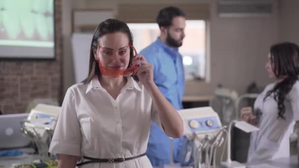 Mladá krásná zubařka si nasazuje ochranné brýle a zapíná vytvrzovací světla, zatímco její mnohonárodnostní kolegové hovoří v pozadí. Kavkazský stomatolog pózující s vybavením. — Stock video