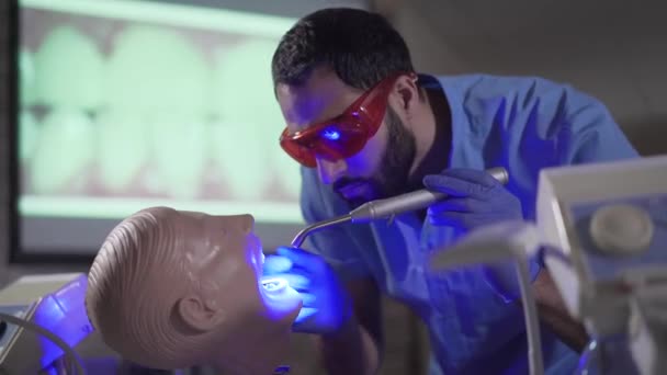 Retrato del ocupado asistente estomatólogo de Oriente Medio practicando con el maniquí dental. Joven enfocado en gafas protectoras usando luces de curado. Profesión, medicina, estomatología . — Vídeos de Stock