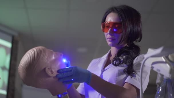 Concentré jeune femme brune en utilisant des lumières de durcissement sur mannequin dentaire. Portrait de stomatologue caucasien occupé dans les lunettes de protection pratiquant la dentisterie. Profession, mode de vie, qualification . — Video