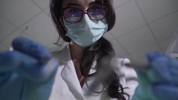 Extrema face close-up da bela mulher branca morena em óculos e máscara protetora usando equipamentos odontológicos na câmera. Retrato de jovem dentista com instrumentos. Odontologia . — Vídeo de Stock