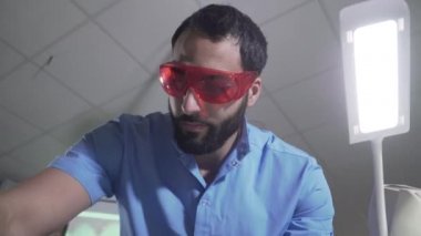 Mavi üniformalı ve koruyucu gözlüklü ciddi bir Orta Doğulu hasta ziyareti için hazırlanıyor. Ofisteki yakışıklı sakallı erkek dişçinin portresi. Sağlık, ilaç, meslek.