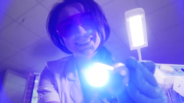Patientens synvinkel av leende tandläkare med hjälp av härdande ljus. Vacker ung kaukasisk kvinna i skyddande glasögon och handskar gör tandmanipulation. Stomatologi, medicin. — Stockvideo