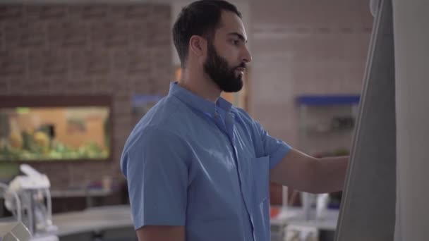 Vue latérale d'un bel homme du Moyen-Orient confiant écrivant à bord dans le public de la dentisterie. stomatologue professionnel enseignant aux étudiants. Mode de vie, éducation, médecine, odontologie . — Video