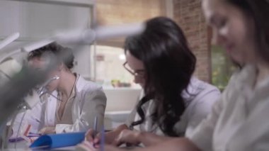 Beyaz üniformalı beyaz dişçi öğrencilerinin stomatoloji üzerine yazı yazma testleri. Ciddi kadınlar dişçiler arasında masada oturuyor. Eğitim, öğrenme, tıp.