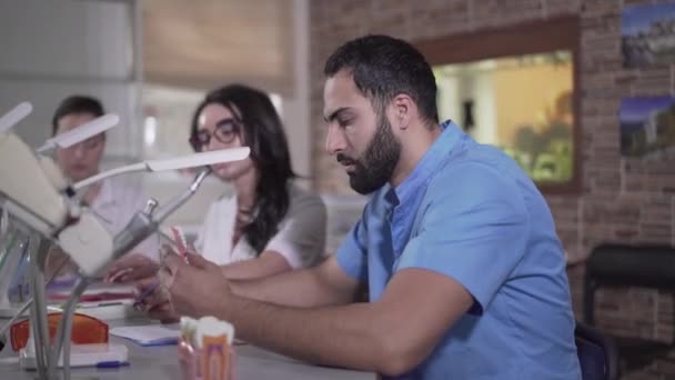 Vue latérale de l'homme épuisé du Moyen-Orient regardant les dents lancées et soupirant. Étudiant en dentisterie masculine fatigué étudiant en stomatologie avec des camarades de groupe. Médecine, éducation, profession .