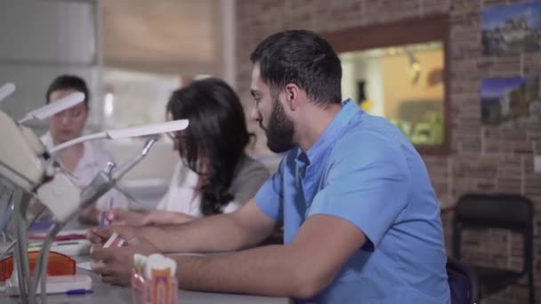 Primo piano di stanco studente di odontoiatria mediorientale che prende la testa con le mani. Vista laterale del giovane in uniforme blu che sospira tra il pubblico dentale. Istruzione, sovraccarico, esaurimento, medicina . — Video Stock