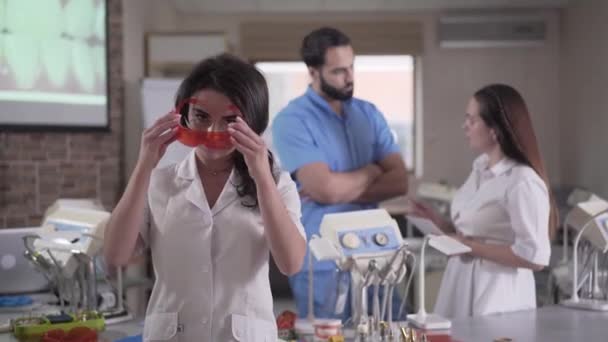 Retrato de jovem morena colocando óculos de proteção e sorrindo para a câmera. Dentista feminina caucasiana encantadora em uniforme posando dentro de casa. Medicina, profissão, ocupação . — Vídeo de Stock