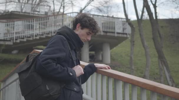 Vue latérale du garçon caucasien triste avec sac à dos debout sur les escaliers dans le parc de la ville et la pensée. Malheureux étudiant masculin qui passe la journée d'automne à l'extérieur. Solitude, tristesse, dépression, mode de vie . — Video