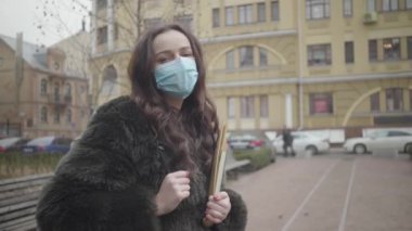 Şehir caddesinde elinde kitaplarla duran yüzü maskeli güzel esmer kadın portresi. Salgın sırasında koruyucu maske takan bilinçli bir vatandaş. Covid-19, sağlık, ilaç, karantina.