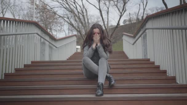 도시 공원의 계단에 앉아 울고 있는 백인 소녀의 모습. 아름다운 갈색 머리 여자에게 문제가 있어요. 슬픔, 우울증, 슬픔, 스트레스. — 비디오