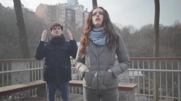 Retrato de la hermosa morena joven mirando hacia otro lado como novio nervioso regañándola desde el fondo. Novio gritándole a bonita novia caucásica en el parque de otoño. Conflicto, ruptura . — Vídeos de Stock