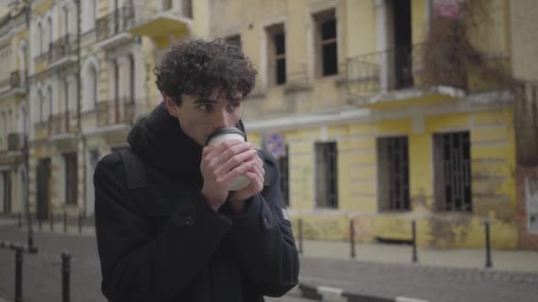 Ung brunett vit pojke med lockigt hår tittar runt i främmande stad och dricker kaffe. Positiv manlig turist reser utomlands på höstdagen. Fritid, livsstil, turism. — Stockvideo